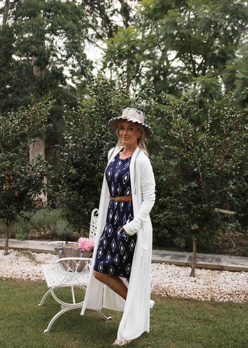 Middle aged woman wearing a white long-line pocket cardigan over a patterned midi dress & white flat sandals (Product image)