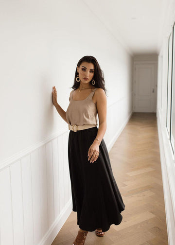 A woman posing in a hallway wearing a bronze sleeveless cami top, black maxi skirt and a pair of brown high heel sandal