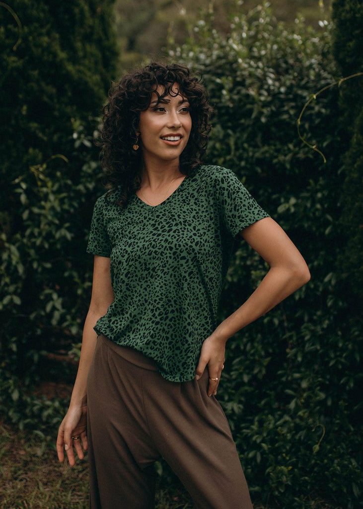 An outdoor image of a woman wearing a green leopard printed top and brown relaxed pants