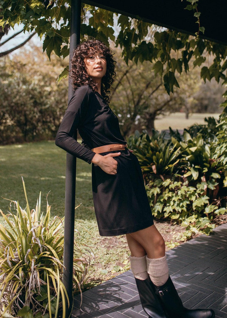 An outdoor shot of a woman wearing a black long sleeve dress with brown belt and a pair of black boots