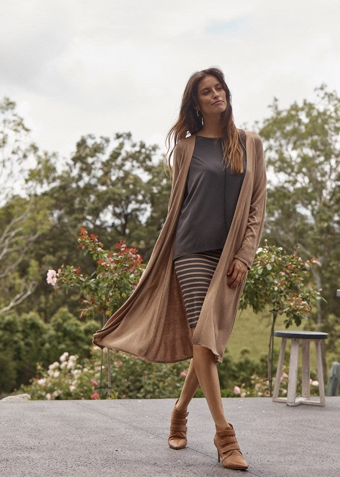 An outdoor shot of a woman wearing a brown bamboo dusty jacket, grey top and a striped black and brown midi skirt and a pair of brown pointed ankle boots