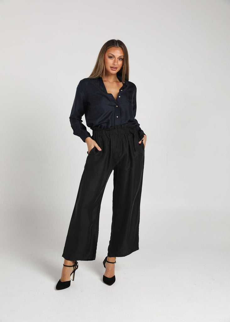 A woman posing while both of her hands are insider her black pant's pockets whil e wearing a black button down long sleeve and black pointed high heels