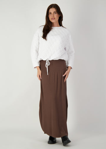 A woman standing in a grey backdrop while wearing a white pullover, brown ribbed bamboo maxi skirt and a pair of black boots (Product image)