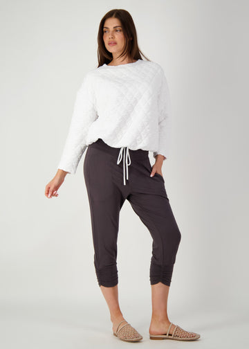 A woman posing confidently in a grey backdrop while wearing a white pullover, brown bamboo cropped pants and a flat brown sandals (Product image)