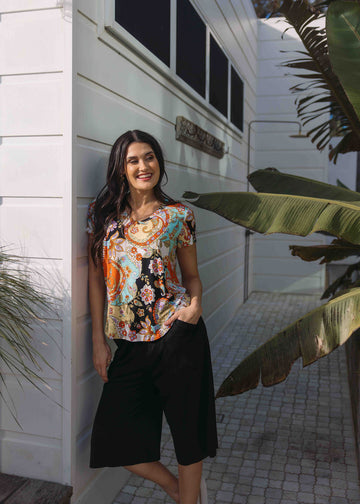 An outdoor shot of a woman smiling confidently while wearing a printed bamboo v-neck shirt and black shorts (Product image)